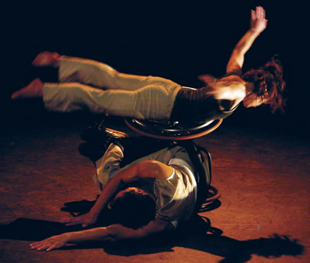 Dois bailarinos da AXIS Dance Company executam uma elevação; um deles utiliza cadeira de rodas.